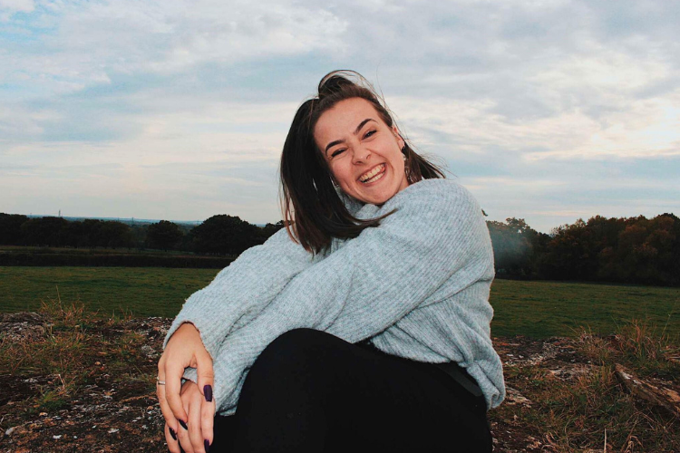 Image: Sophie Smith smiling, sat in a field with trees in the background, wearing a grey jumper and black jeans.