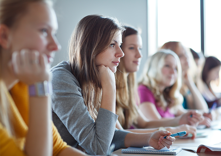 Image: A line of students at a long desk, focusing on a young female with long brown hair and her head leaning on her hand